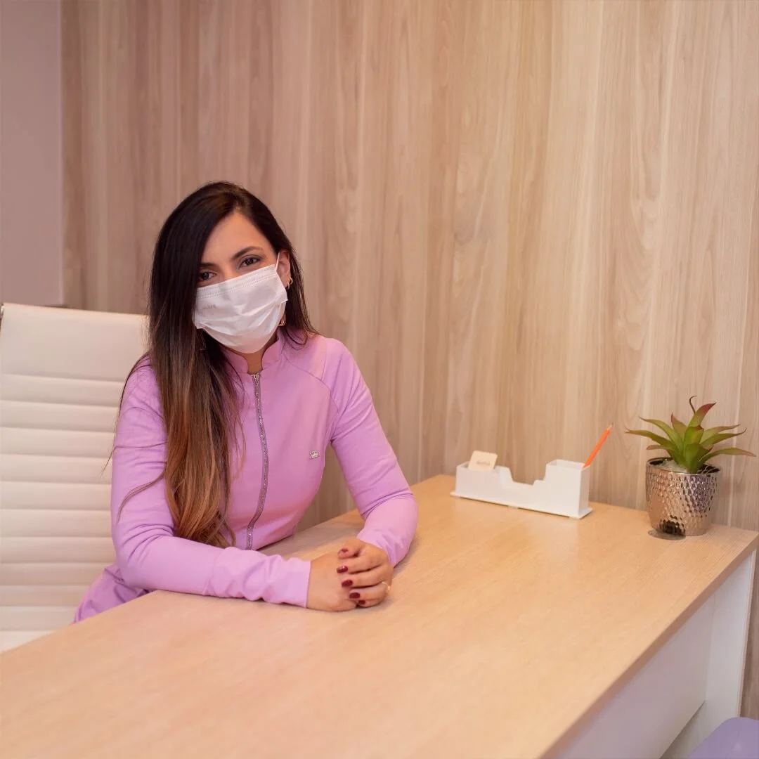 Doutora Luciana D'Angelo, dentista em Ouro Preto, sorrindo, de máscara, com os braços sobre a mesa e as mãos entrelaçadas.